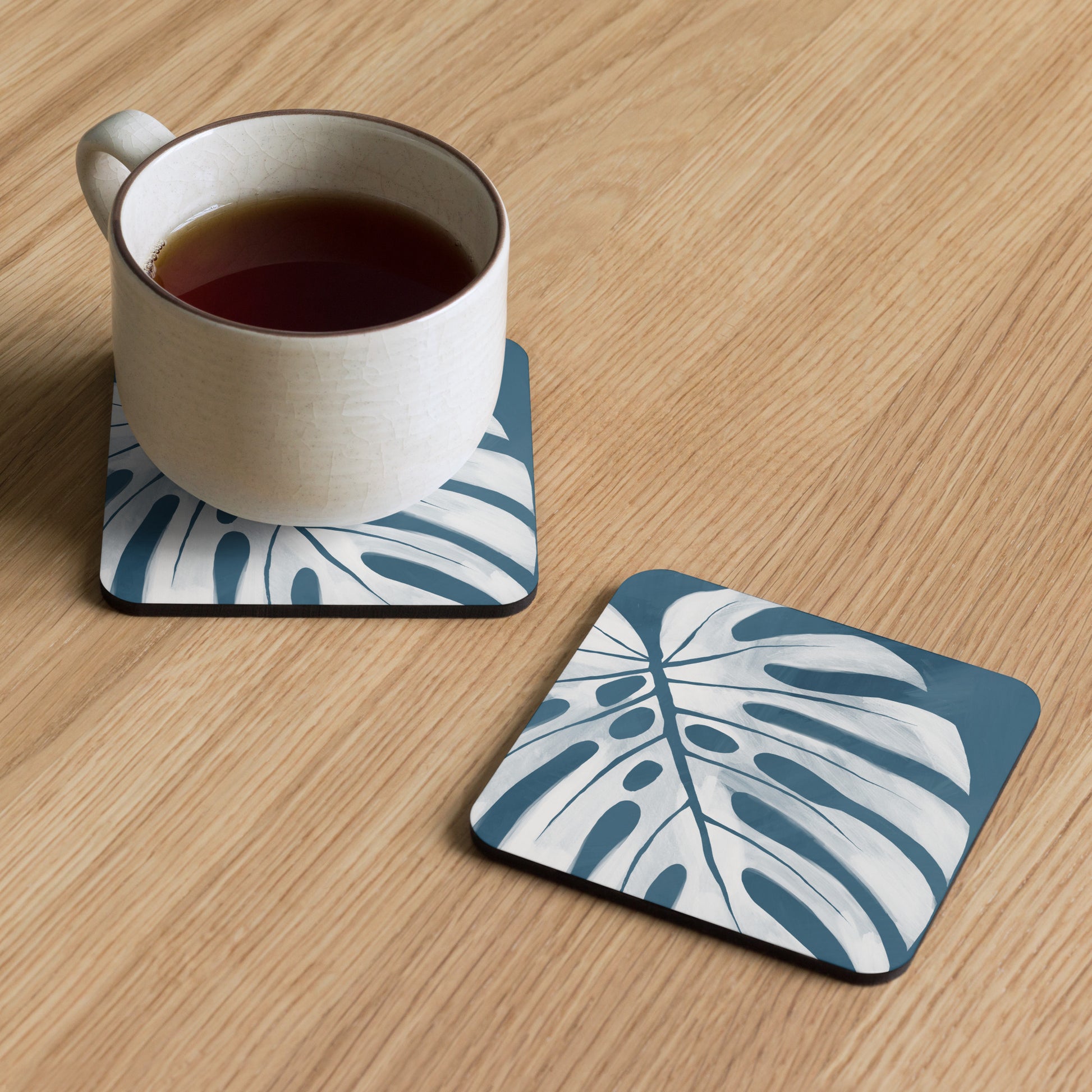 A coffee cups sits on a coaster atop a pine table.  Next to it, a bare coaster with a blue background and a printed white monstera leaf.